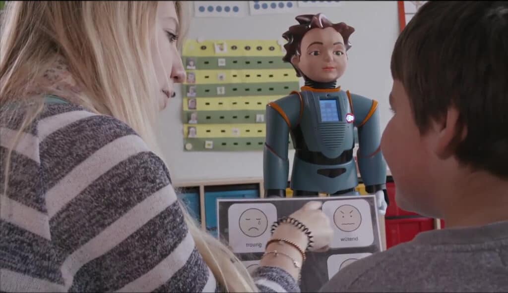 Een vrouw en een jongetje kijken naar een kaart met gezichtjes met verschillende emoties. Op de achtergond staat een robot, wiens emotie ze proberen af te lezen.