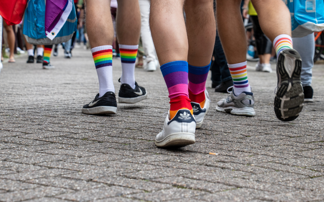 In beeld zie je de blote kuiten van mensen met sokken in regenboogkleuren op straat