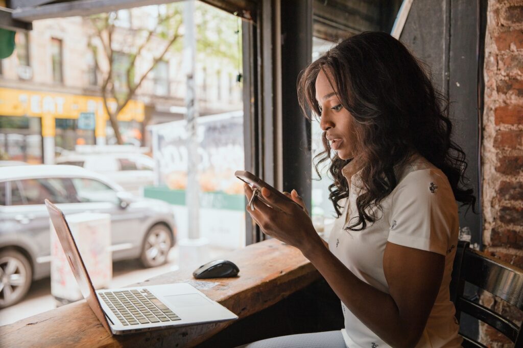 Een vrouw kijkt op haar telefoon terwijl ze achter haar laptop zit in een koffietentje