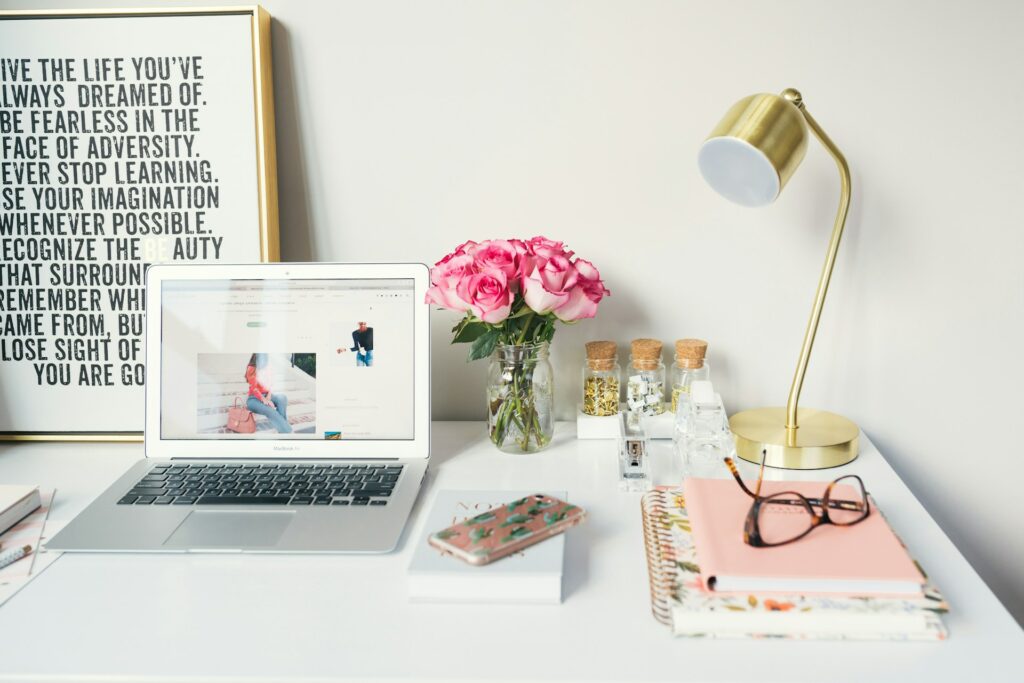 een laptop op een wit bureau met roze rozen, een gouden lamp en wat notitieblokken