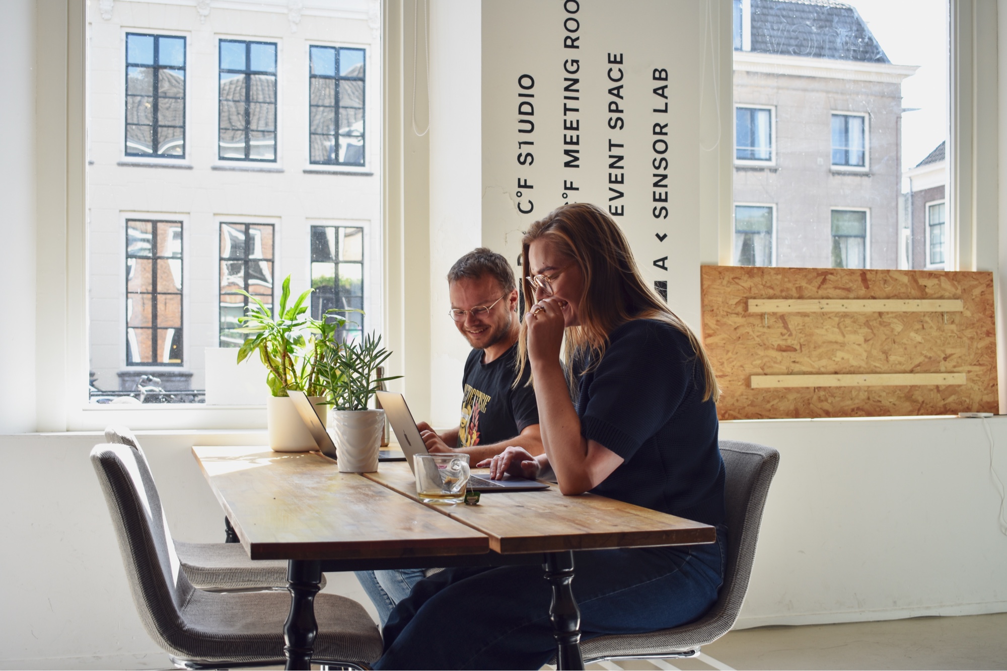 Pim en Emma zitten aan een tafel in het kantoor van digitaal toegankelijk bij een raam. Ze lachen allebei.