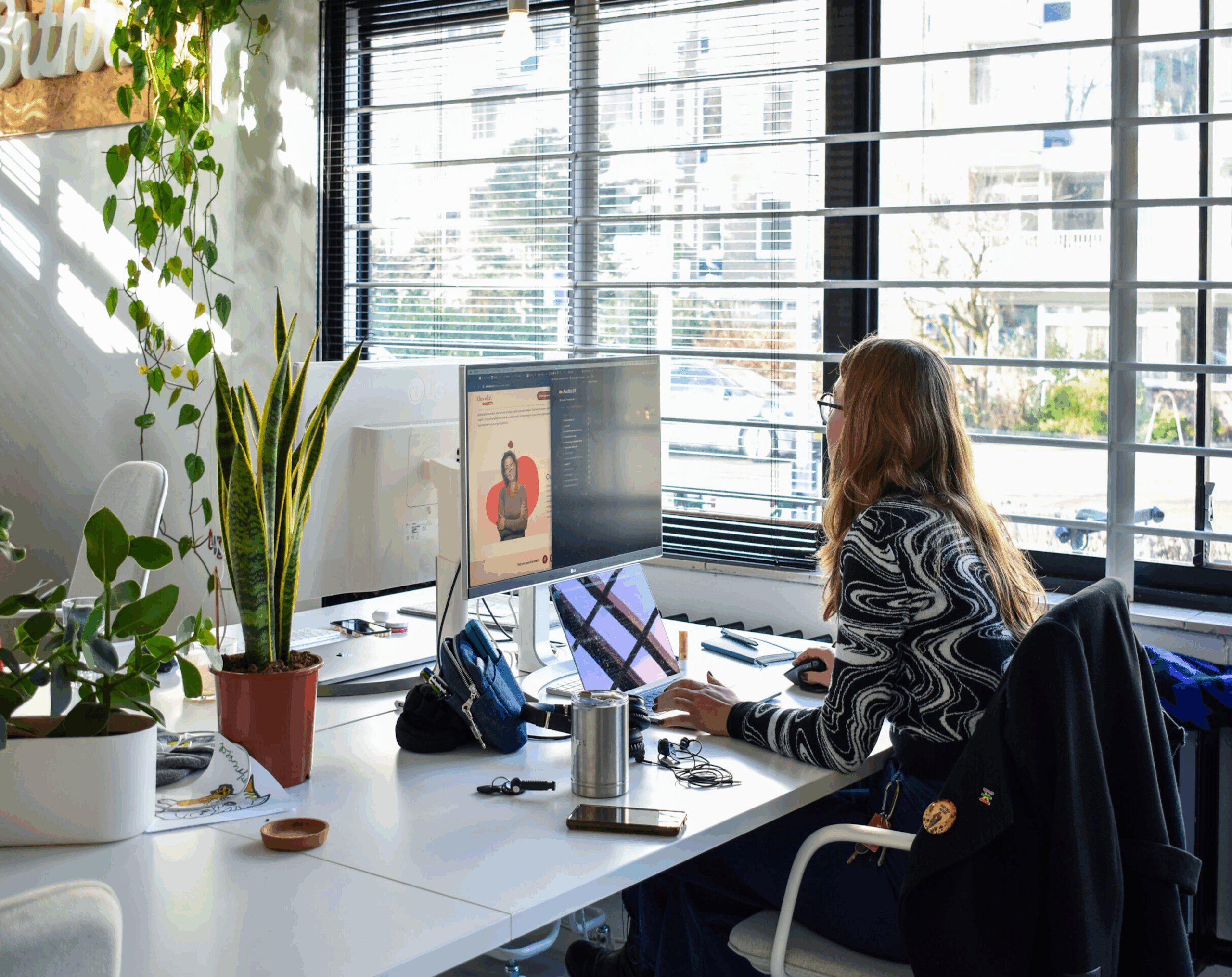 Foto van de beneden verdieping. Dit is nog een foto van het aude kantoor en toont een wit bureau en iemand die voor haar laptop zit te werken nast een groot raam waar een luxaflex voor zit
