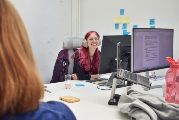 Marieke zit achter haar computer met een koptelefoon op en lacht naar Emma