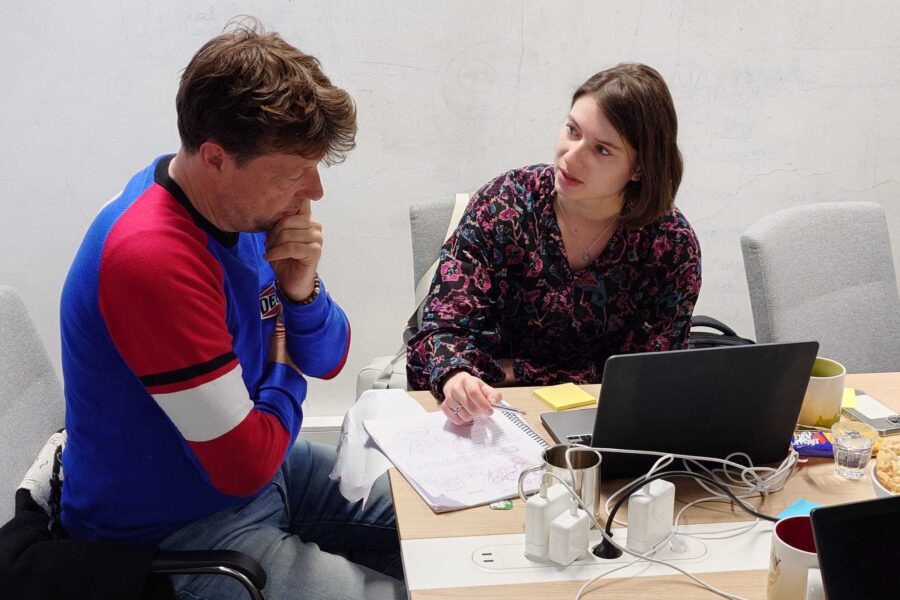 Een man en een vrouw zitten in een ruimte achter een laptop. De vrouw legt iets uit aan de man