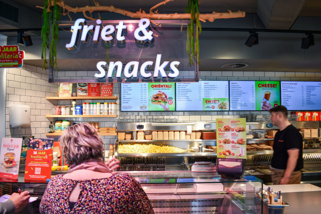 Een oudere vrouw staat voor de kassa bij Kwalitaria. Op de achtergrond zie je de woorden friet & snacks hangen