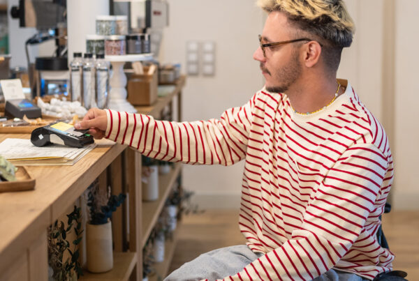 Foto van een aantrekkelijke man in een rolstoel die met zijn pinpas een betaling doet in een hippe koffietent. Hij heeft een vrolijke streepjestrui aan.