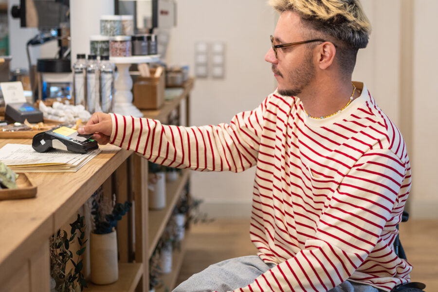 Foto van een aantrekkelijke man in een rolstoel die met zijn pinpas een betaling doet in een hippe koffietent. Hij heeft een vrolijke streepjestrui aan.