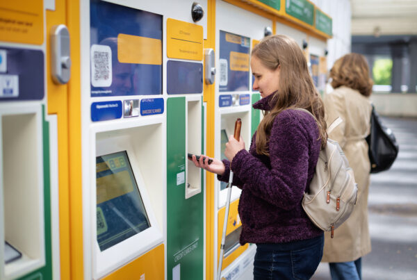 Een vrouw is met haar telefoon bij een kaartjeautomaat een treinkaartje aan het kopen. Ze heeft een rugzak op en houdt een geleidestok vast.